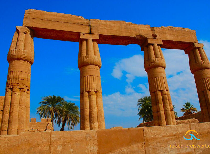 Überreste des Karnak-Tempel. Vier Steinsäulen die oben Steinblöcke tragen. Im Hintergrund Palmen