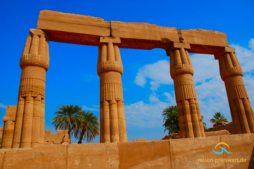 Überreste des Karnak-Tempel. Vier Steinsäulen die oben Steinblöcke tragen. Im Hintergrund Palmen